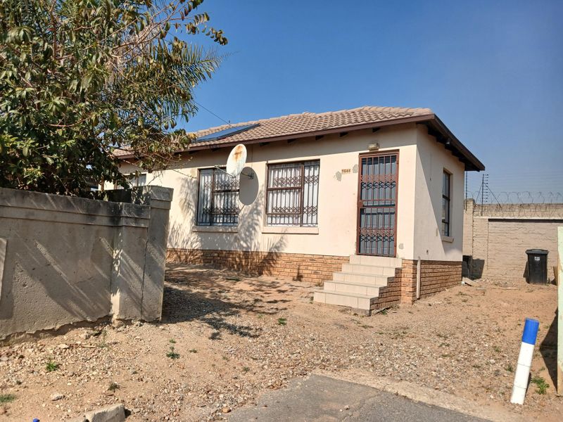 Front elevation of a single-story house featuring a tiled roof, solar water heater, security gates, and a perimeter wall with an unpaved yard area.