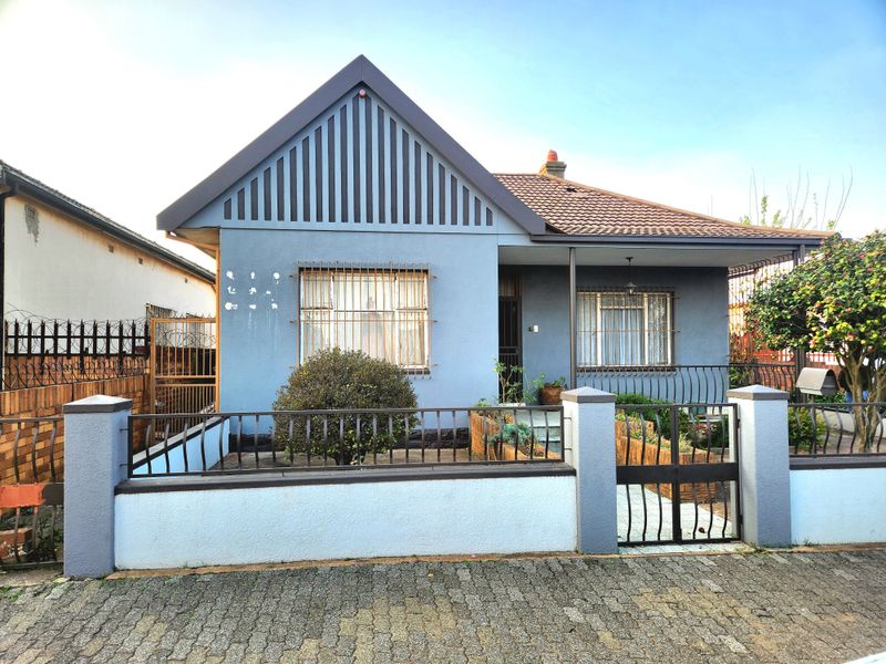 Front elevation of a single-story house featuring a tiled roof, covered porch, and secure perimeter wall with metal gate and security bars.