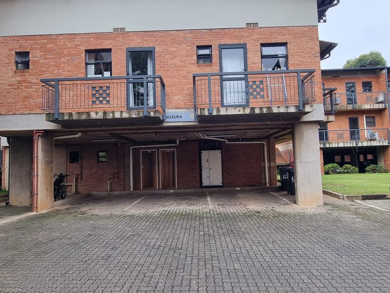 Exterior view of a brick apartment building featuring covered ground-level parking bays and upper-level balconies with metal railings.