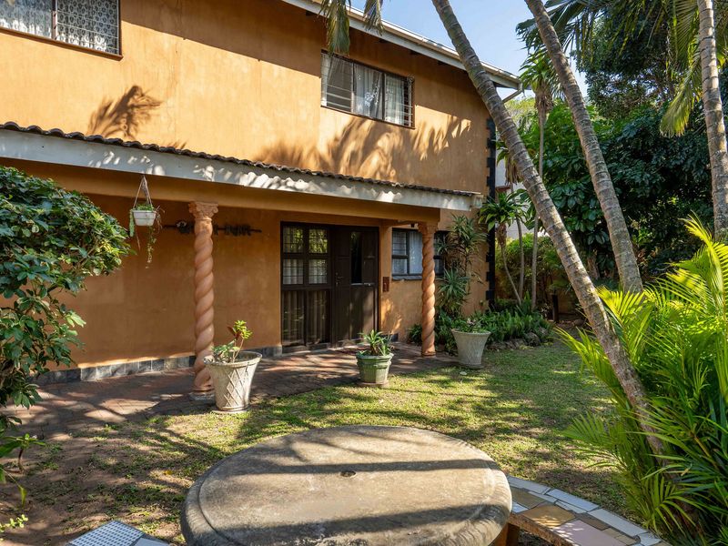 Rear exterior view of the house featuring a covered patio with decorative columns, brick paving, and established garden surroundings.