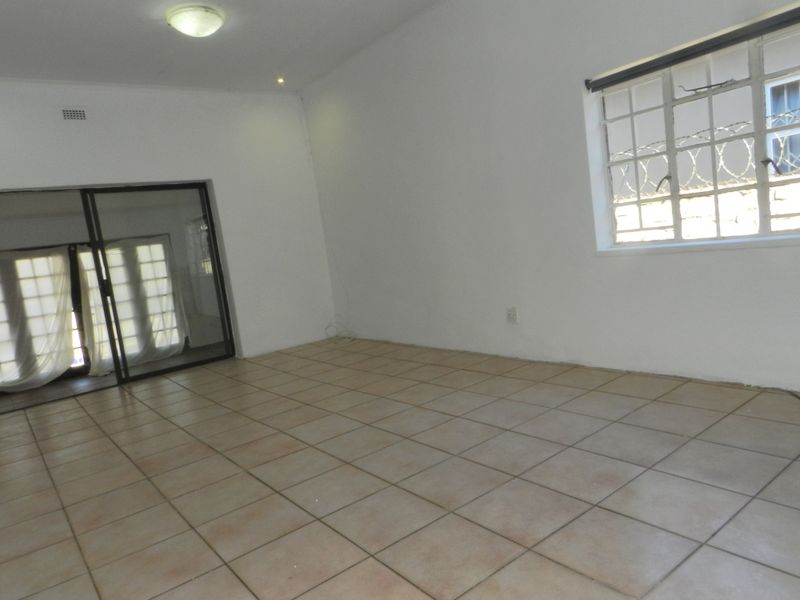 An empty, neutrally painted living room featuring ceramic floor tiles, a large window with security bars, and a sliding glass door leading outside.