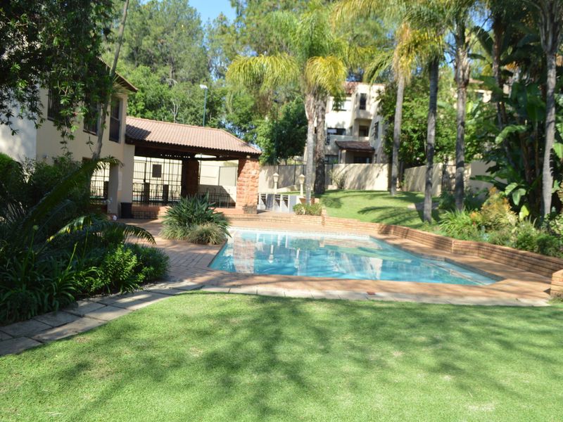 A well-maintained communal swimming pool area featuring brick paving, a covered patio with stone pillars, and established garden landscaping.