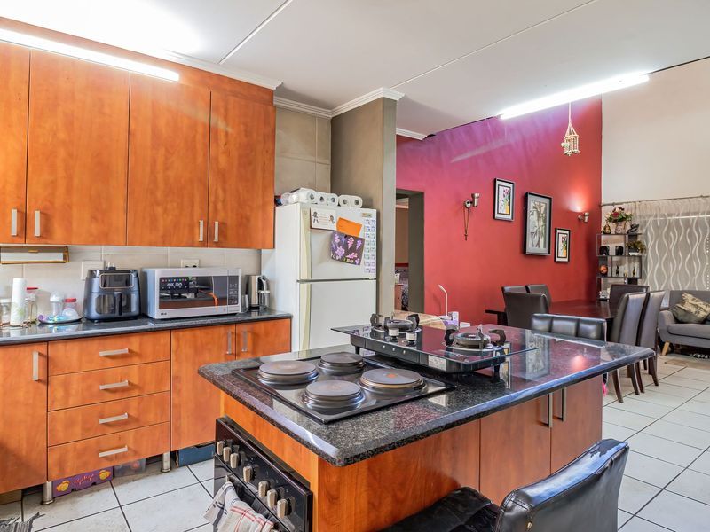 Open-plan kitchen and living area featuring wood-finish cabinetry, granite countertops, a central island with a built-in hob, and tiled flooring throughout.