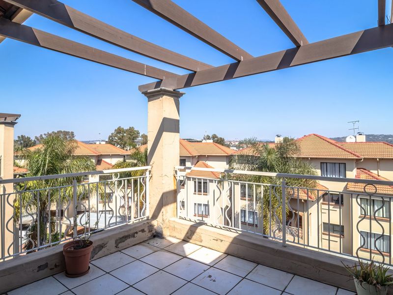 A spacious balcony featuring a wooden pergola, tiled flooring, and wrought iron railings, offering an elevated view of the surrounding apartment complex.