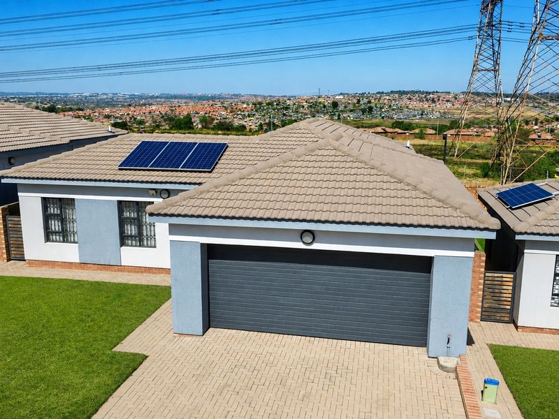 Aerial view of a residential property featuring a tiled roof with solar panels, a paved driveway, a garage, and a well-maintained lawn.