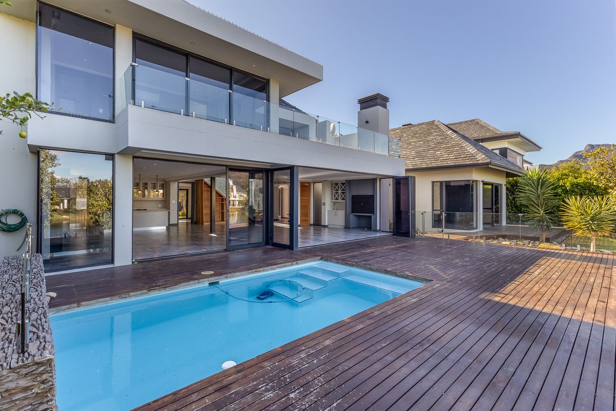 Rear exterior view featuring a swimming pool, timber decking, glass balustrades, and a covered patio area with a built-in braai.