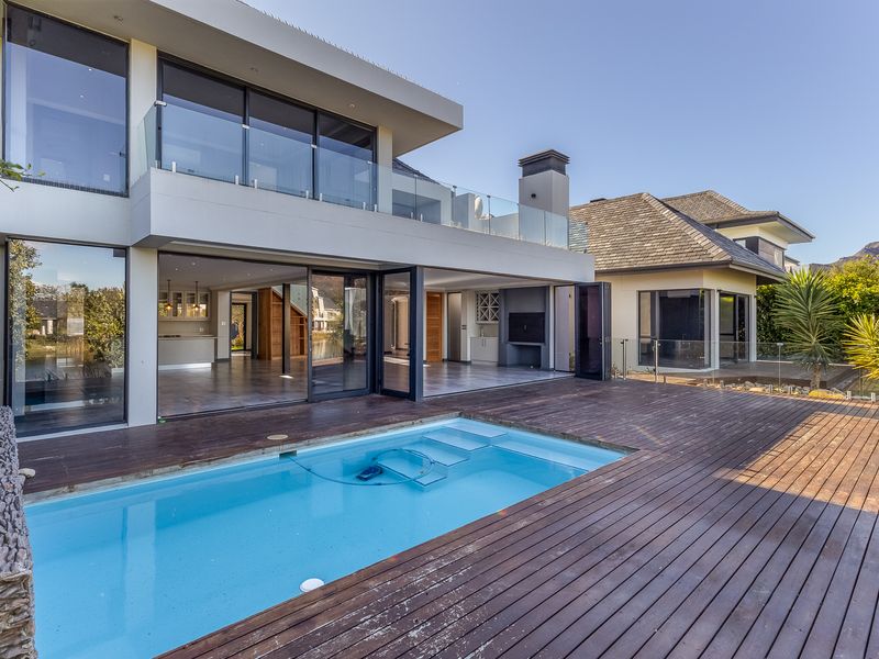 Rear exterior view featuring a swimming pool, timber decking, glass balustrades, and a covered patio area with a built-in braai.