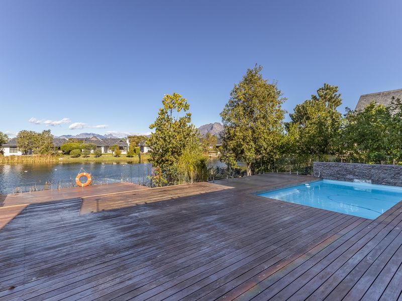 Outdoor pool area featuring extensive timber decking, a glass balustrade, and a stone feature wall overlooking a scenic lake.