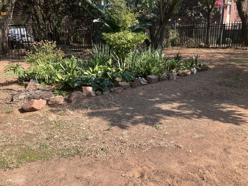A private garden area featuring established trees, a landscaped plant bed with stone edging, and secure metal perimeter fencing.