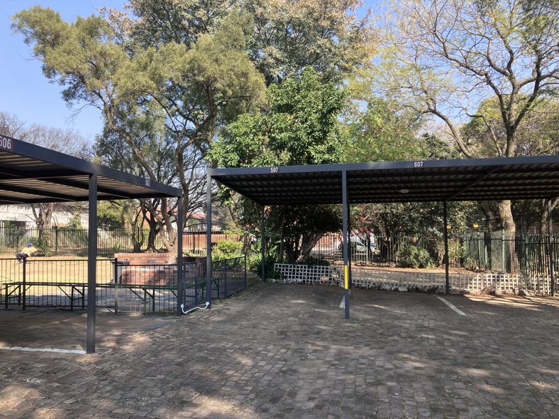 Outdoor parking area featuring covered carports with numbered bays and a built-in brick braai facility within a fenced complex.