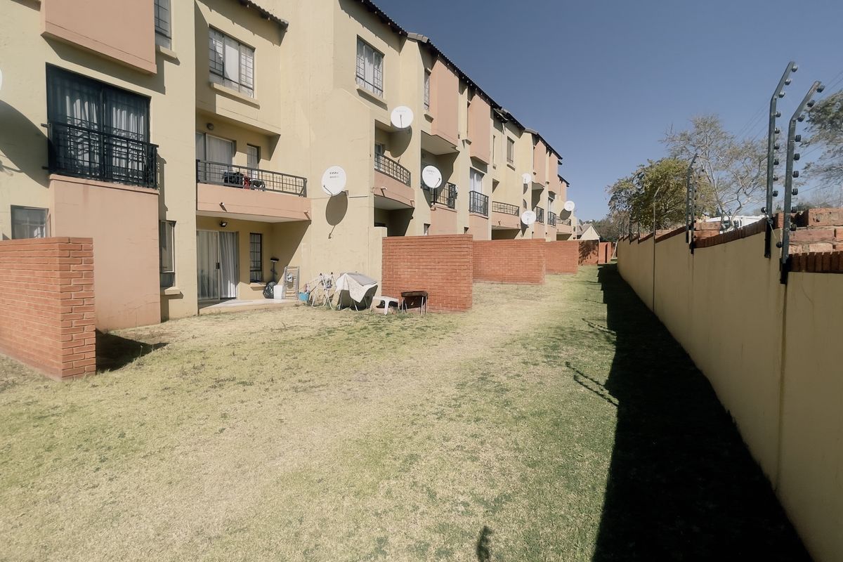 Rear view of a multi-story apartment complex featuring private brick-walled garden sections, balconies, and a perimeter wall with electric security fencing.