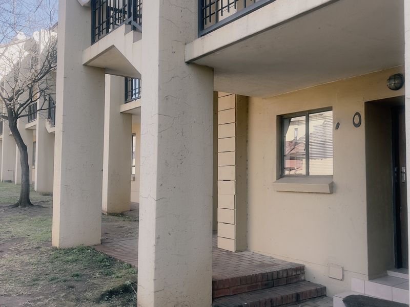 Exterior view of an apartment building entrance featuring concrete pillars, brick paving, and metal balcony railings with visible signs of exterior wear.