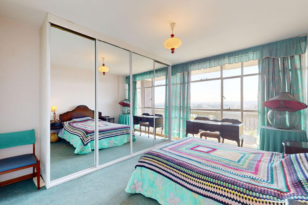 A spacious bedroom featuring a large sliding mirror wardrobe, wall-to-wall carpeting, and a large window providing natural light and exterior views.