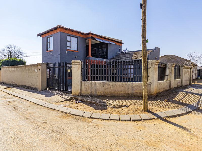 Two-story residential property featuring a grey facade with orange accents, enclosed by a perimeter wall with integrated metal security fencing.