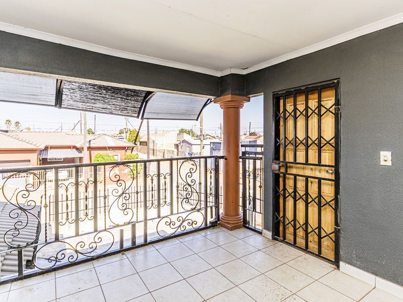A covered balcony featuring tiled flooring, a decorative column, and a wrought iron balustrade, providing an outdoor space with security gate access.