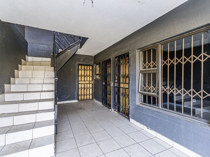 Covered outdoor patio area featuring tiled stairs, metal security gates, and security bars on windows, providing access to the building entrance.