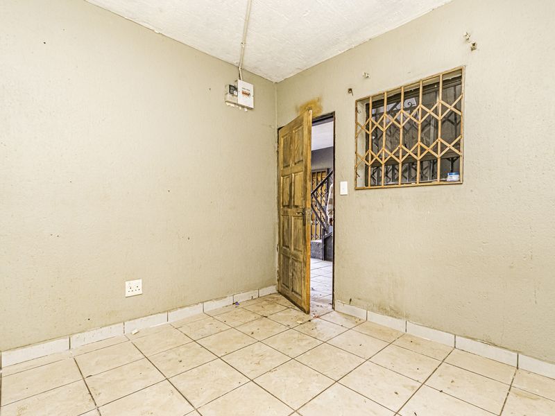An empty, unfinished room featuring tiled flooring, a timber door, and a security-grilled window, requiring cosmetic updates and maintenance.