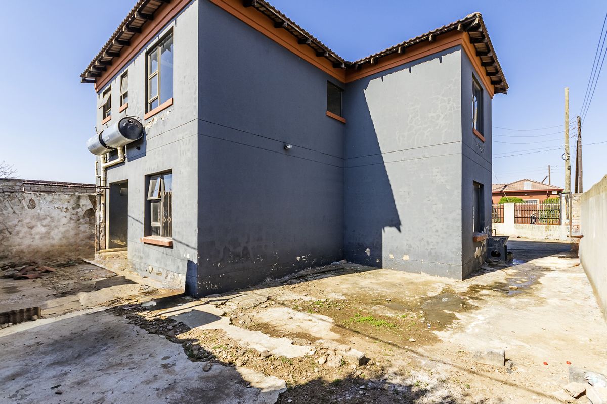 Rear view of a two-story house featuring a solar water heater, tiled roof, and significant wear to the exterior walls and surrounding concrete paving.