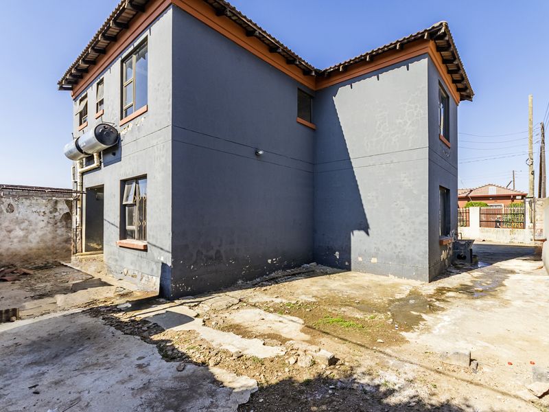 Rear view of a two-story house featuring a solar water heater, tiled roof, and significant wear to the exterior walls and surrounding concrete paving.