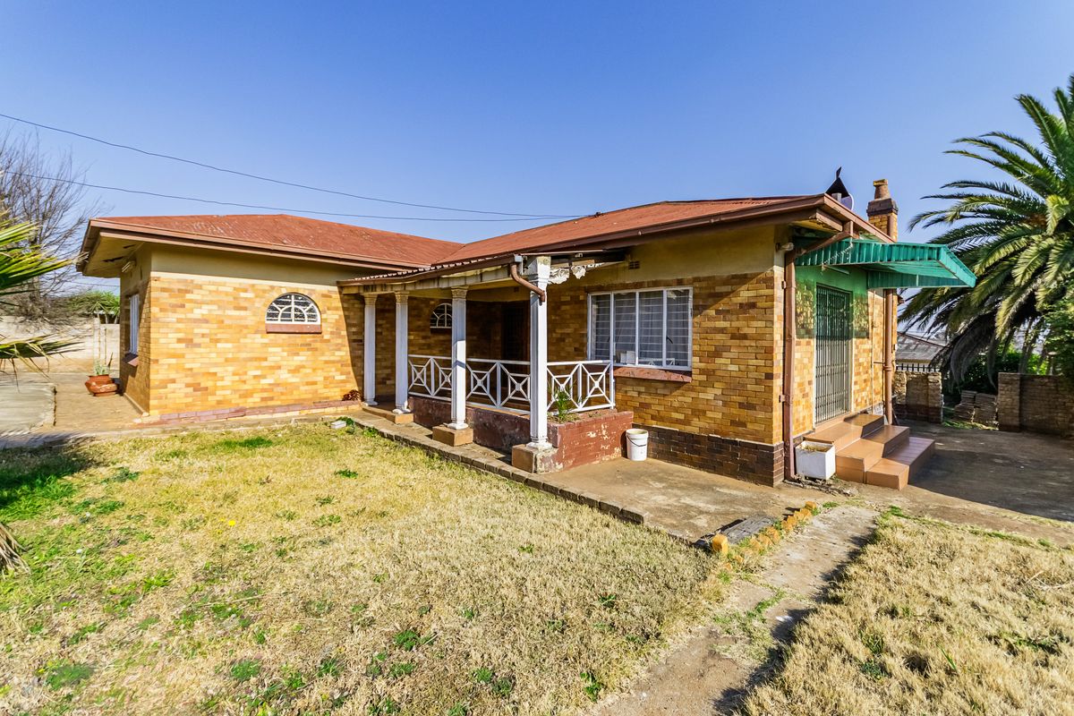 Rear view of a single-story brick house featuring a covered patio, security bars on windows, and a corrugated metal roof.