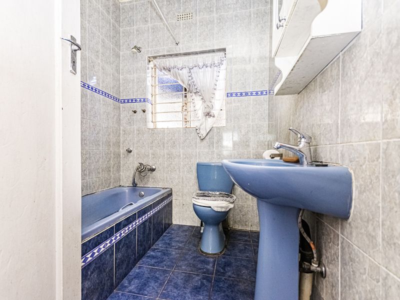 A dated bathroom featuring blue sanitaryware, including a bathtub, pedestal sink, and toilet, complemented by blue and grey ceramic wall and floor tiling.