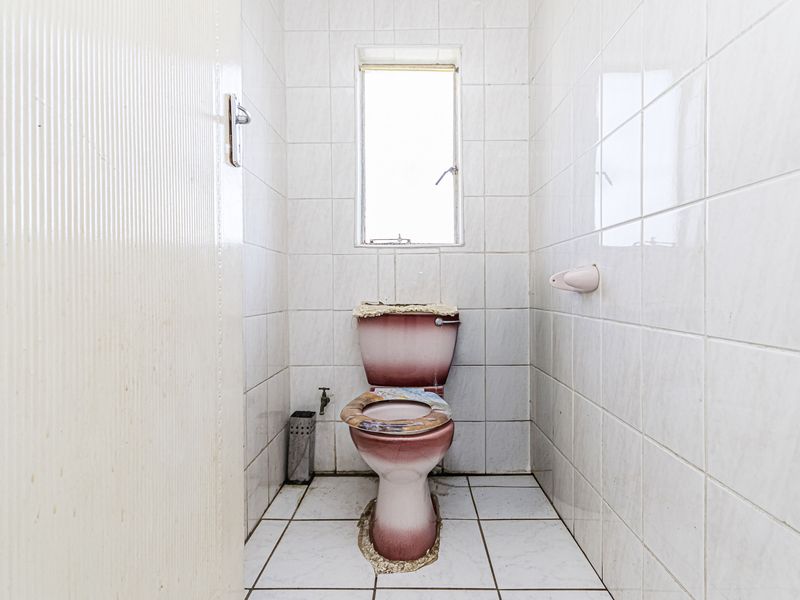 A small, dated bathroom featuring white wall and floor tiles, a window, and a pink toilet suite in need of replacement.