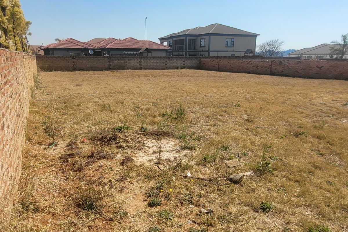 A vacant residential plot enclosed by perimeter brick walls, featuring dry grass and surrounding suburban residential properties.