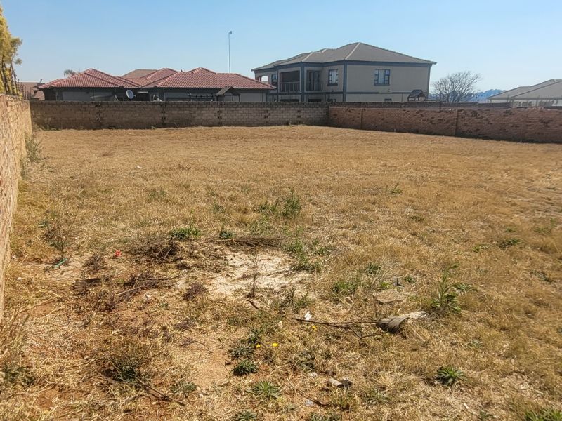 A vacant residential plot enclosed by perimeter brick walls, featuring dry grass and surrounding suburban residential properties.