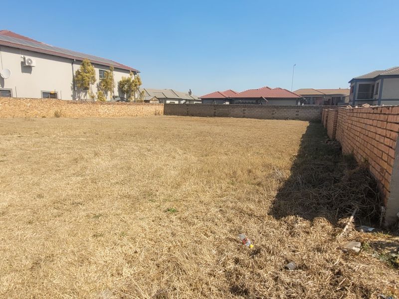 A vacant residential plot featuring dry grass and enclosed by perimeter brick walls in a suburban neighborhood setting.