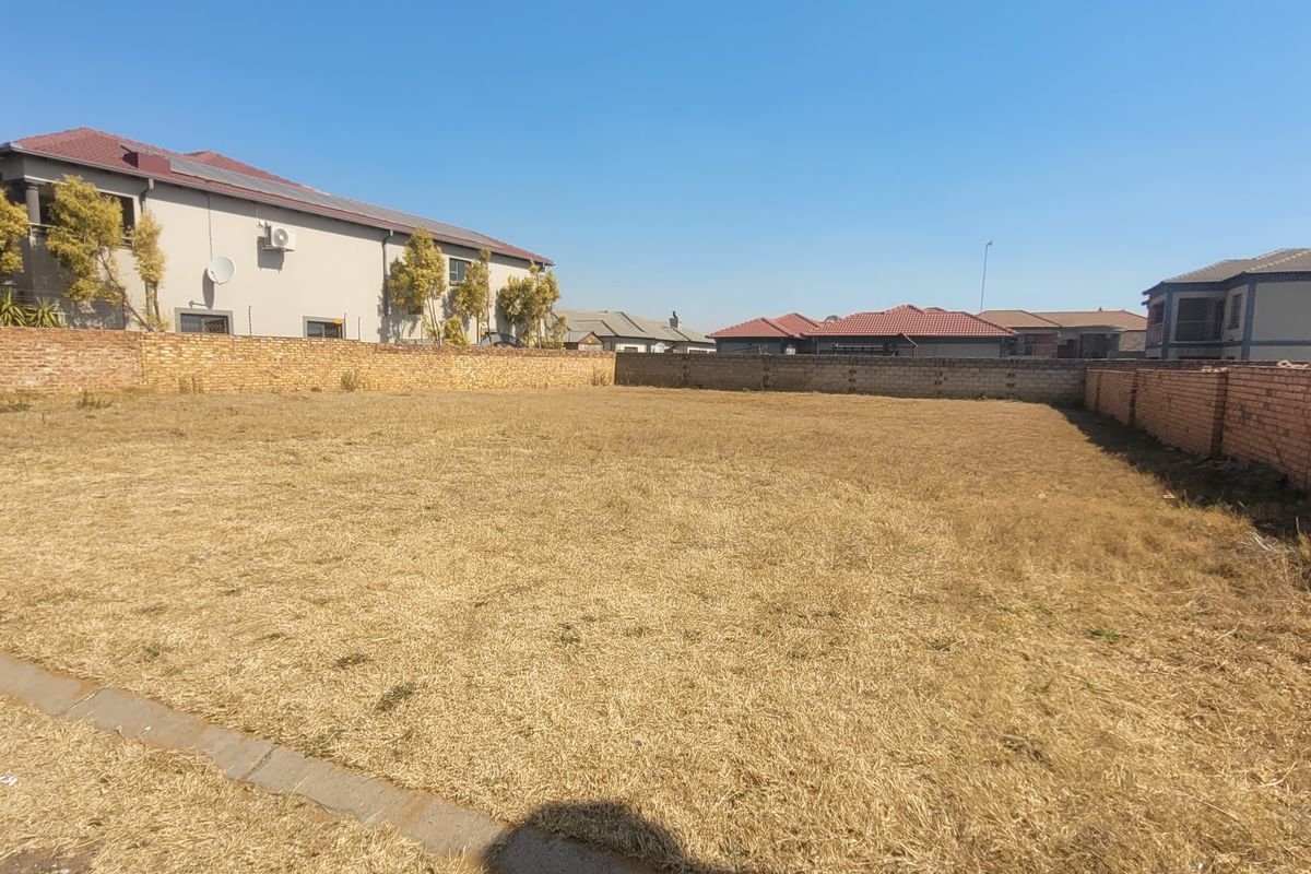 A vacant residential plot featuring dry grass and enclosed by brick boundary walls in a developed suburban neighborhood.