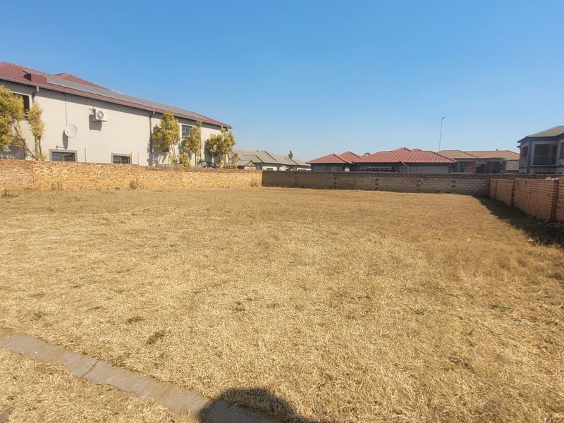 A vacant residential plot featuring dry grass and enclosed by brick boundary walls in a developed suburban neighborhood.