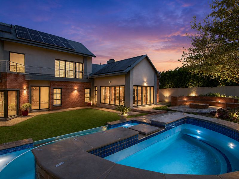 A beautifully landscaped rear exterior featuring a swimming pool, brick and render facade, solar panels, and a spacious patio area at twilight.