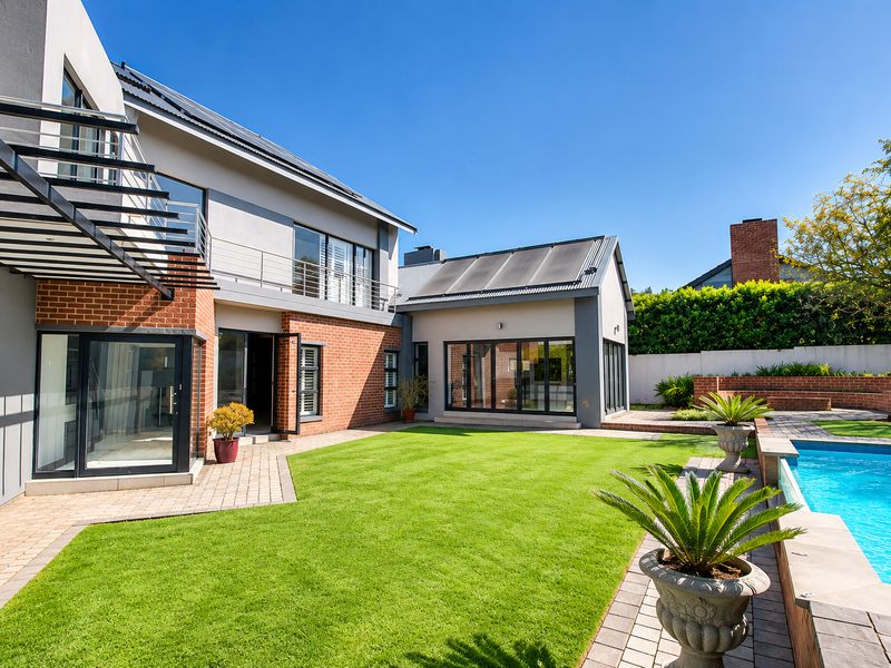 A well-maintained rear exterior featuring a landscaped lawn, paved patio area, swimming pool, and modern architectural details including solar panels and a balcony.