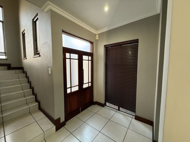 A bright and spacious entrance hall featuring tiled flooring, a wooden double door, and a staircase leading to the upper level.