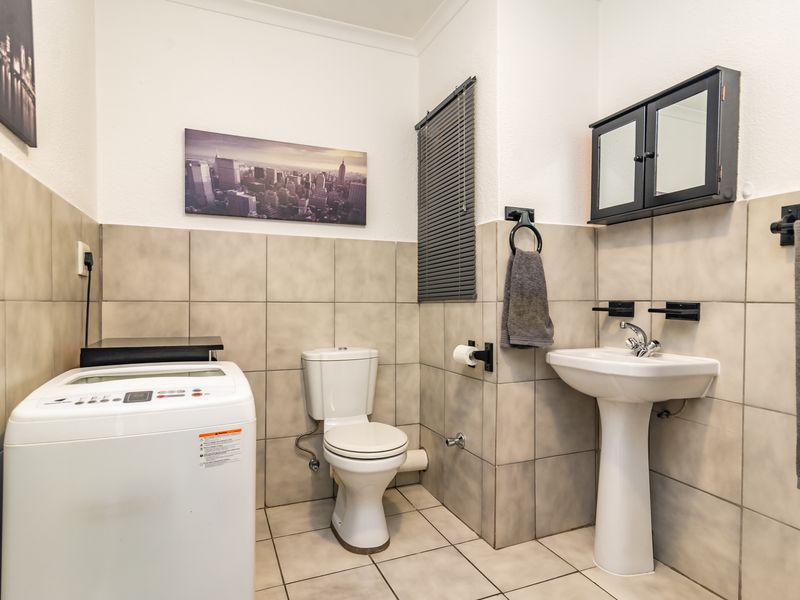 A functional bathroom featuring a pedestal washbasin, toilet, and neutral wall tiling, with space provided for a laundry appliance.