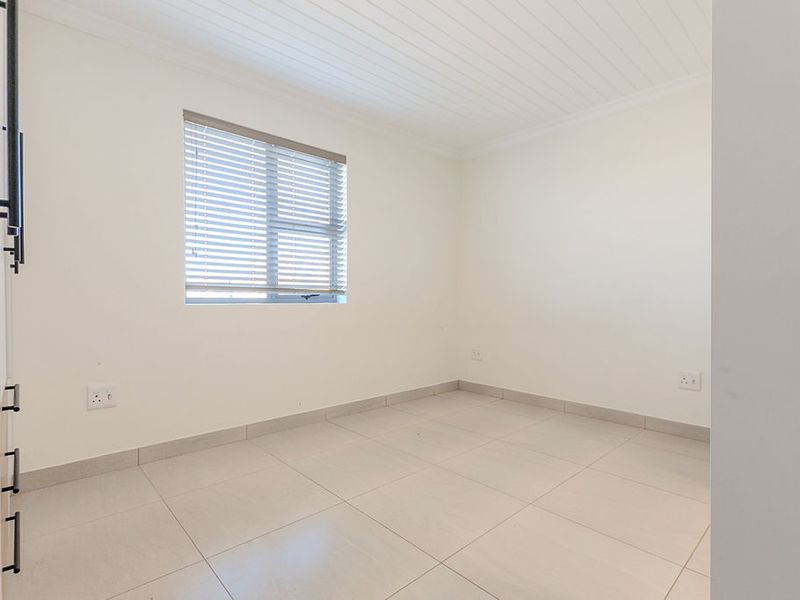A bright, empty bedroom featuring neutral tiled flooring, a tongue and groove ceiling, and a window fitted with venetian blinds.