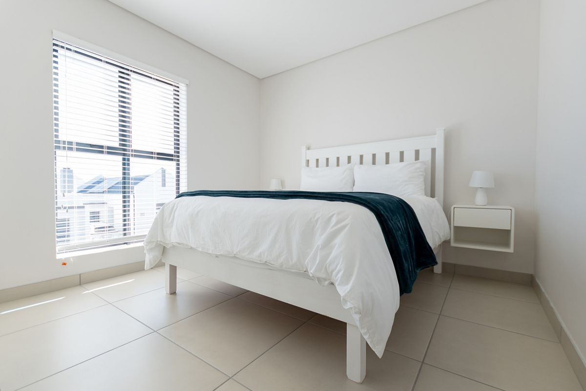 A bright and modern bedroom featuring neutral tiled flooring and large windows fitted with venetian blinds.