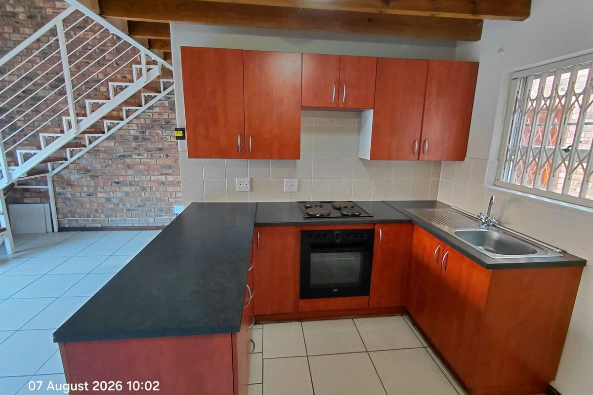 A functional kitchen featuring wooden cabinetry, built-in oven and hob, tiled backsplash, and an adjacent brick feature wall with an open staircase.