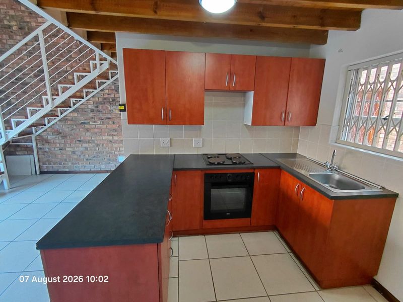 A functional kitchen featuring wooden cabinetry, built-in oven and hob, tiled backsplash, and an adjacent brick feature wall with an open staircase.