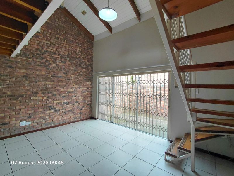 Spacious living area featuring a double-volume vaulted ceiling with exposed timber beams, a striking brick feature wall, tiled flooring, and a staircase.