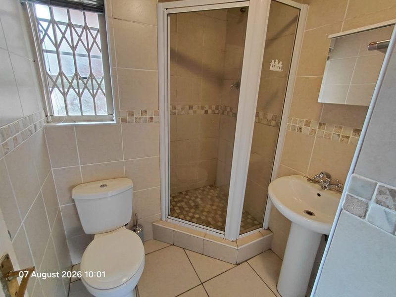 A functional bathroom featuring a corner shower enclosure, pedestal basin, and toilet, finished with neutral wall and floor tiling.