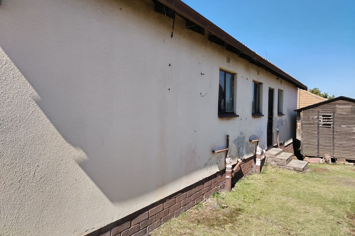 Rear exterior view of the property showing plastered walls with brick plinth, metal window frames, and exposed plumbing infrastructure.