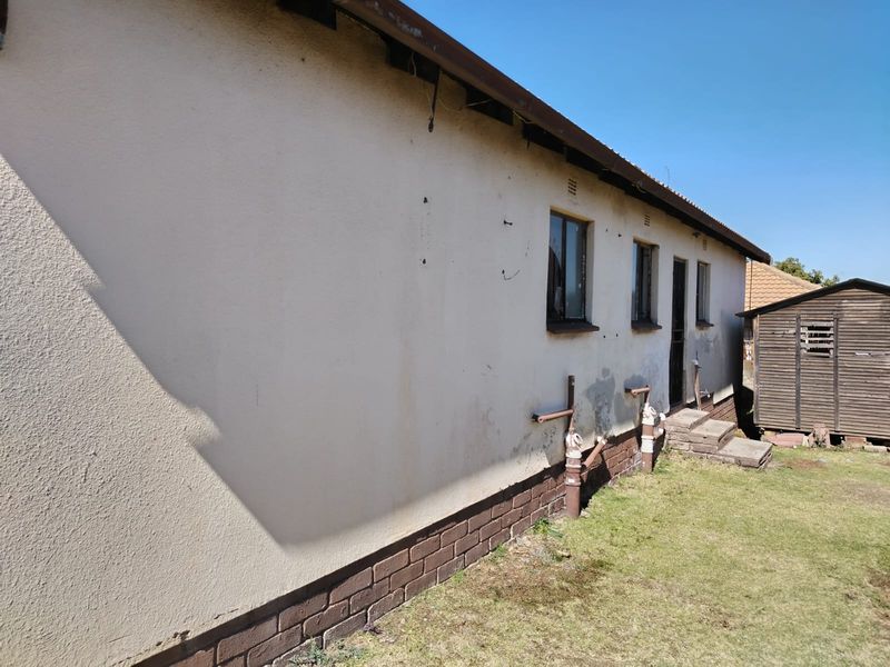 Rear exterior view of the property showing plastered walls with brick plinth, metal window frames, and exposed plumbing infrastructure.
