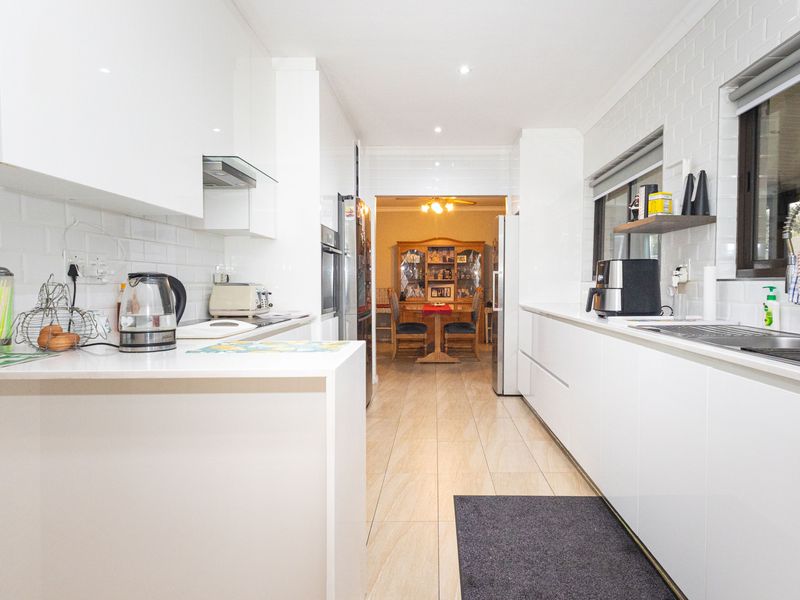 Modern galley-style kitchen featuring white high-gloss cabinetry, subway tile backsplash, integrated appliances, and tiled flooring, leading through to a dining area.