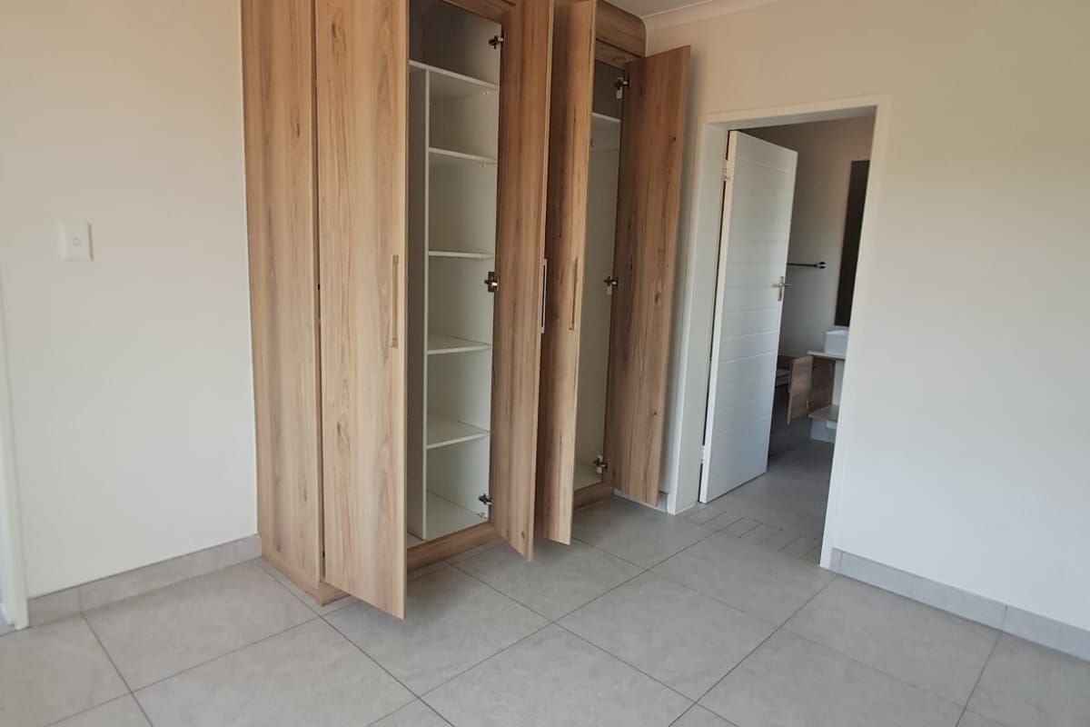 A bright, empty bedroom featuring modern light wood built-in cupboards, neutral tiled flooring, and a clean, minimalist aesthetic.