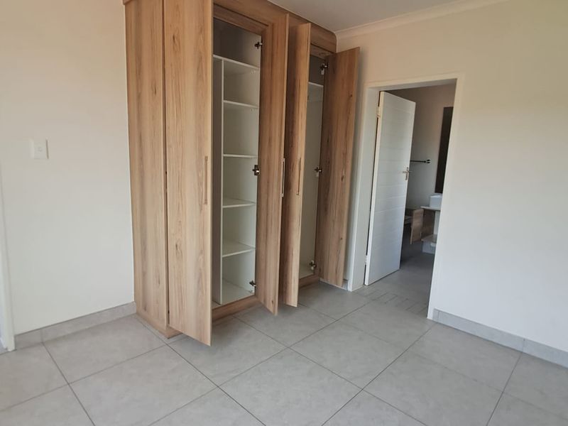 A bright, empty bedroom featuring modern light wood built-in cupboards, neutral tiled flooring, and a clean, minimalist aesthetic.