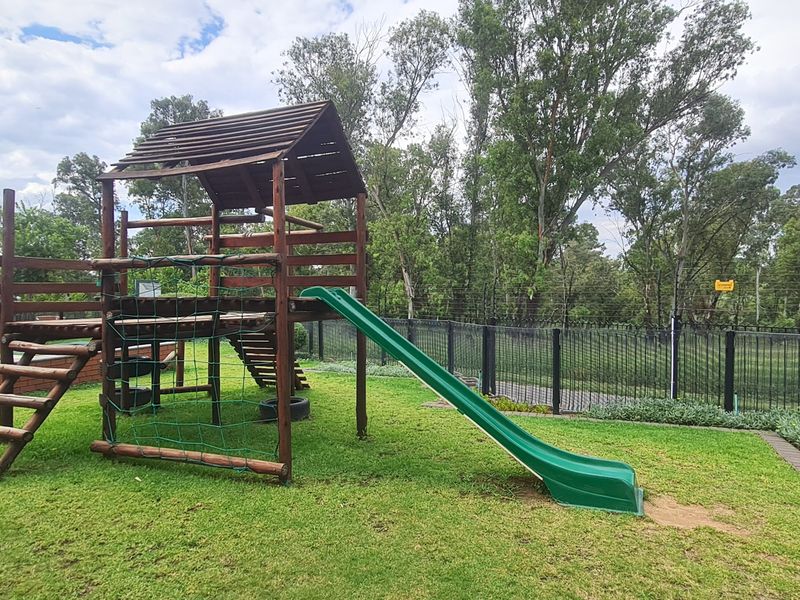 A well-maintained communal garden area featuring a wooden playground structure with a slide, set against a backdrop of mature trees and perimeter fencing.
