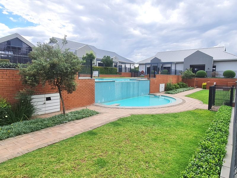 A well-maintained communal pool area featuring a water feature, brick retaining walls, paved pathways, and landscaped garden beds within an apartment complex.