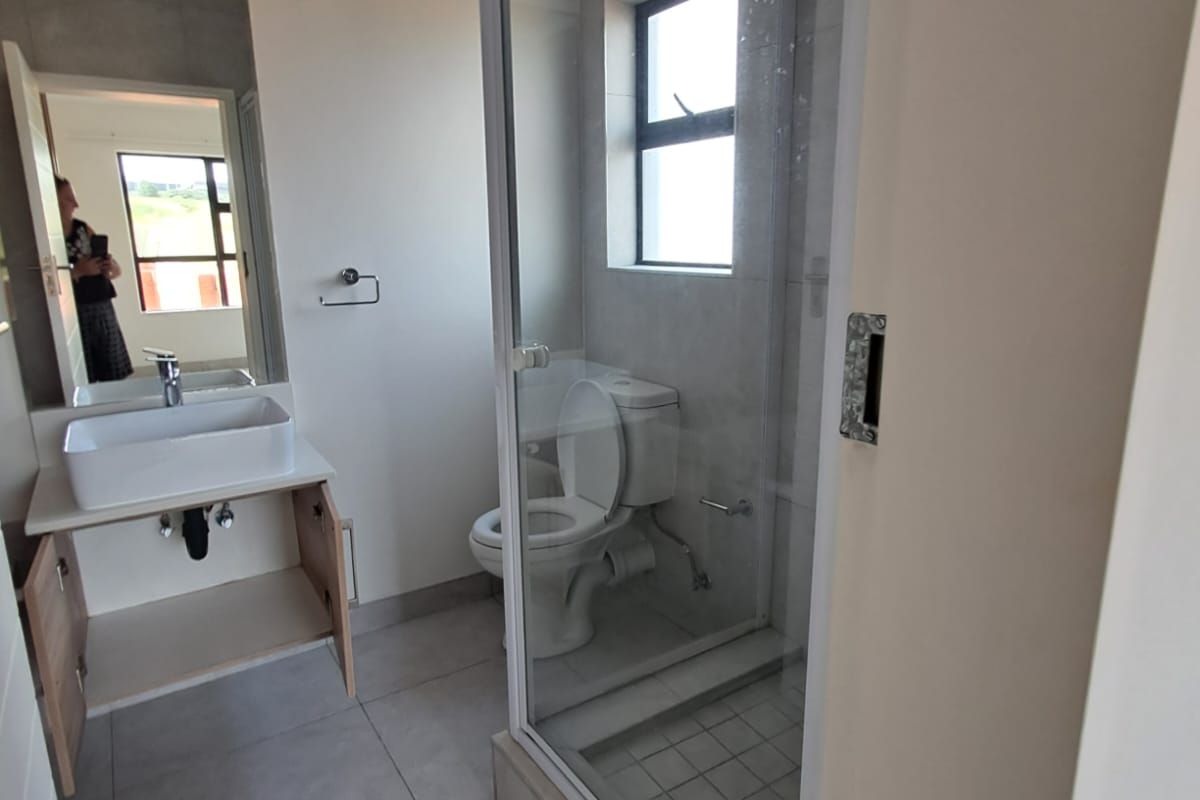 A modern bathroom featuring a glass-enclosed shower, a toilet, and a vanity basin with a large mirror. The space is finished with neutral floor tiles.