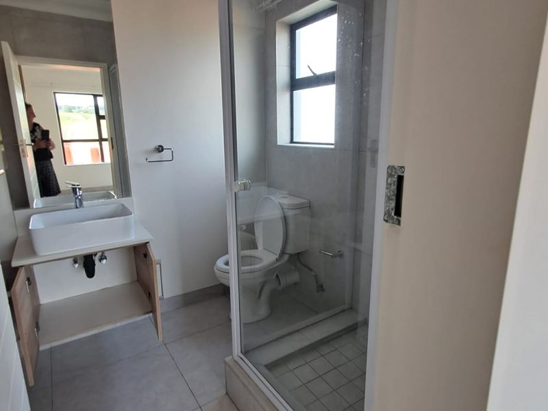 A modern bathroom featuring a glass-enclosed shower, a toilet, and a vanity basin with a large mirror. The space is finished with neutral floor tiles.